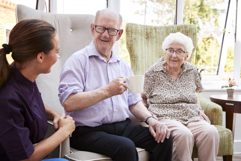 Male And Female Residents Sitting In Chair And Talking With Carer In Retirement Home