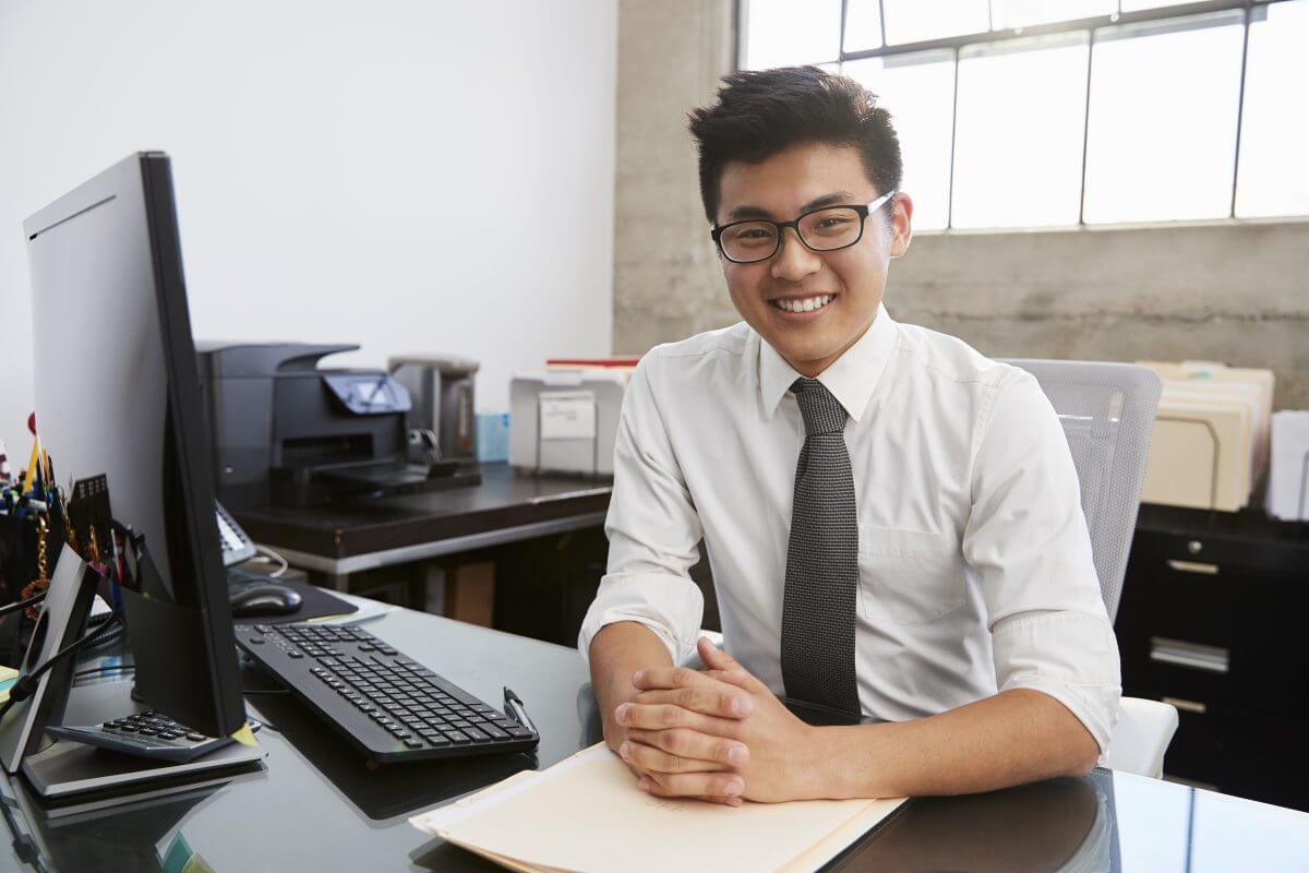 young asian male professional at desk smiling to RU9HW36 e1555930076118